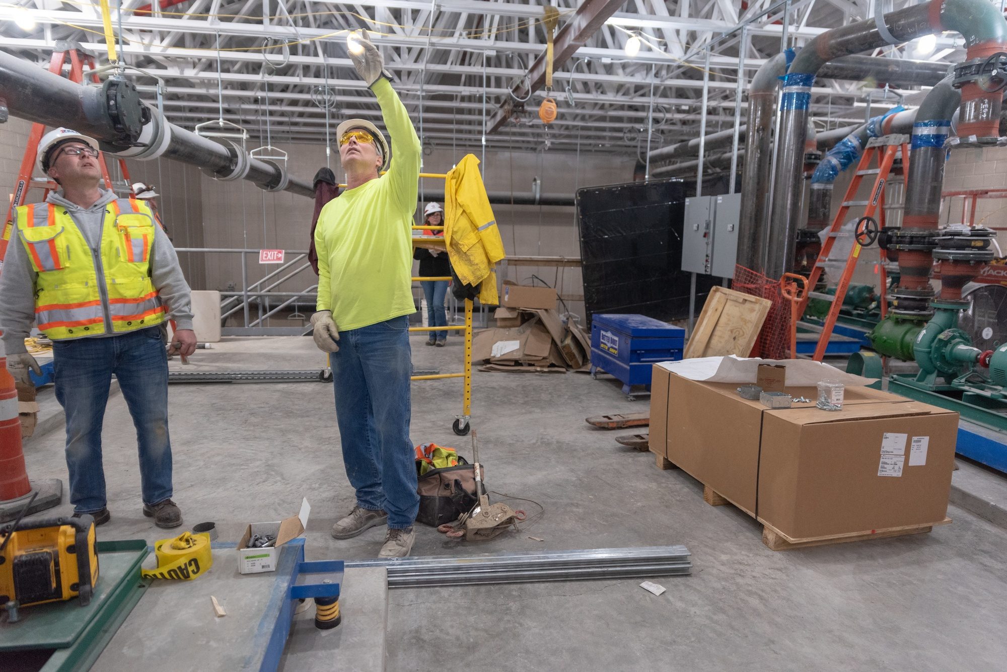 Electrical contractor workers in hard hats and high-visibility clothing reaching up to overhead conduit and hangers inside an unfinished chilled-water plant, with large piping, unopened material crates and tools on the concrete floor