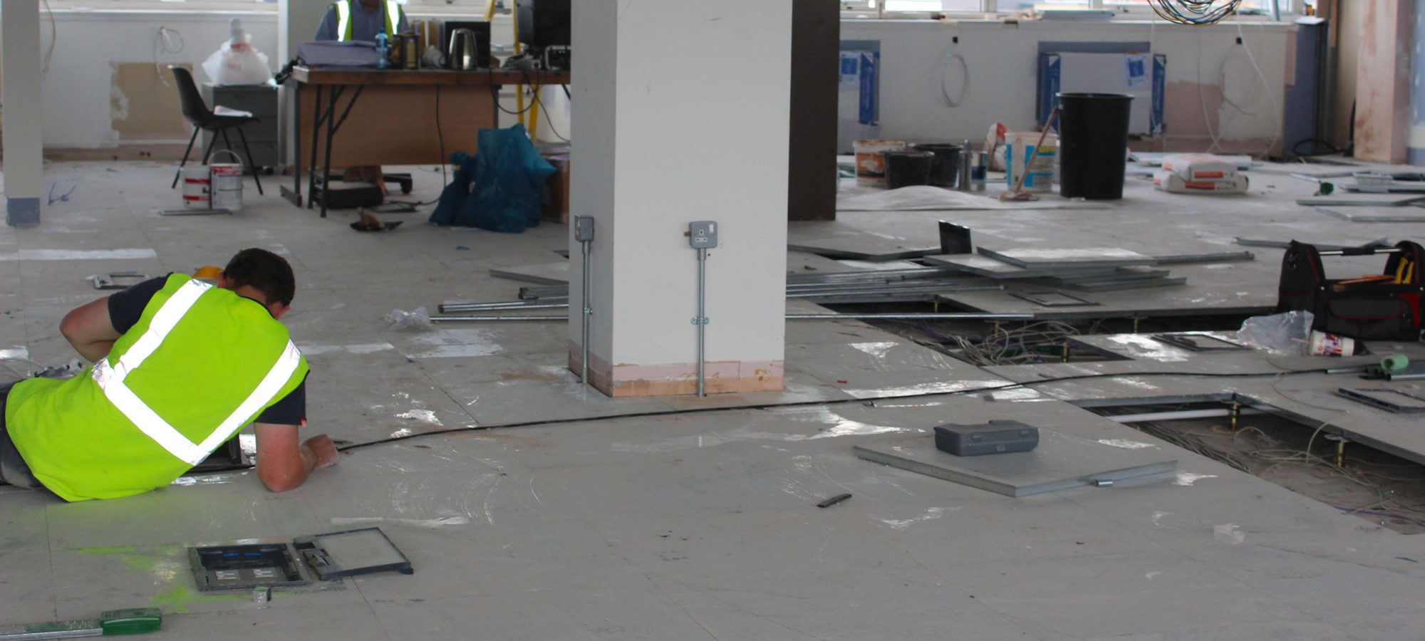 A worker in a high-visibility vest lying on an unfinished floor pulling cable through an open floor box, with exposed cabling, floor panels and tools across the interior fit-out area